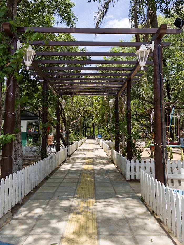 A sunlit pathway under a pergola in an urban park with white picket fences and lanterns.